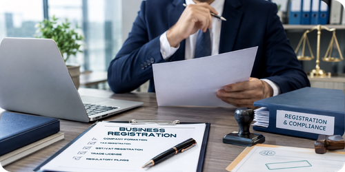 Professional in a suit reviewing a business registration document at a desk with a laptop, stamp, and compliance folder.
